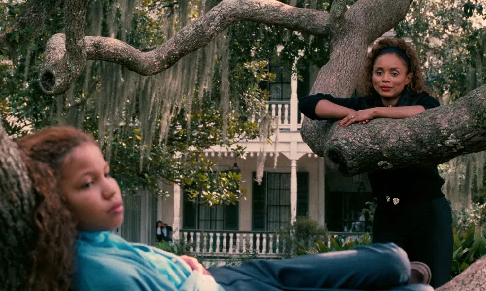 Two women relaxing on large tree branches with a white house and Spanish moss in the background.