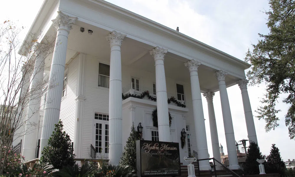 White Greek Revival mansion with tall Corinthian columns and manicured garden in front under a clear sky.