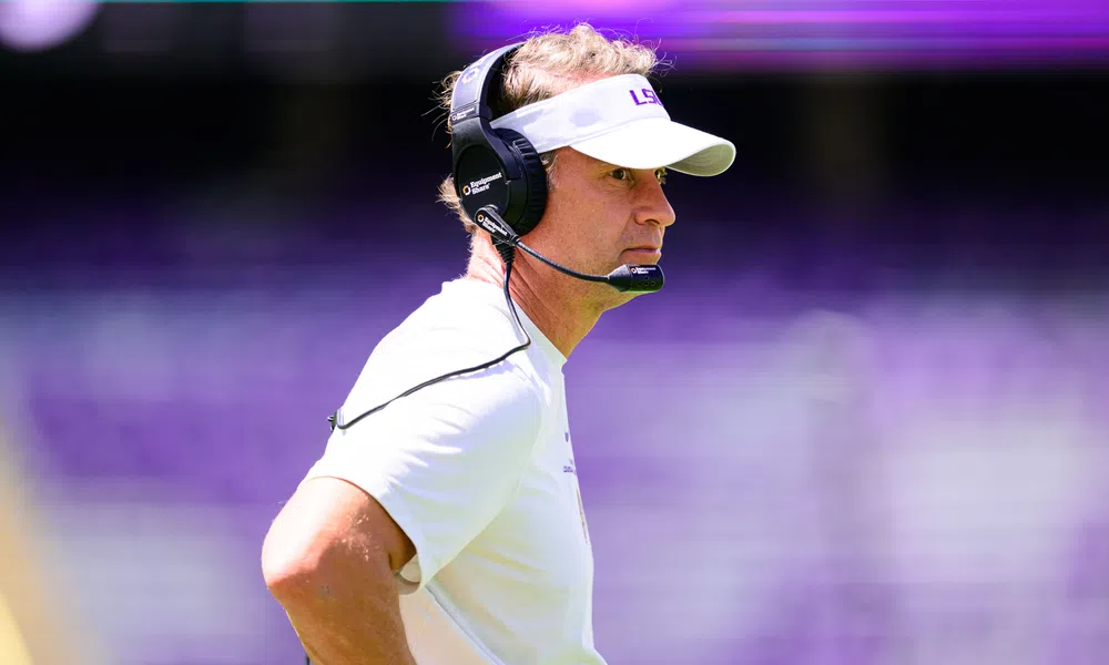 Football coach wearing LSU visor and headset focused during a game with blurred purple stadium background