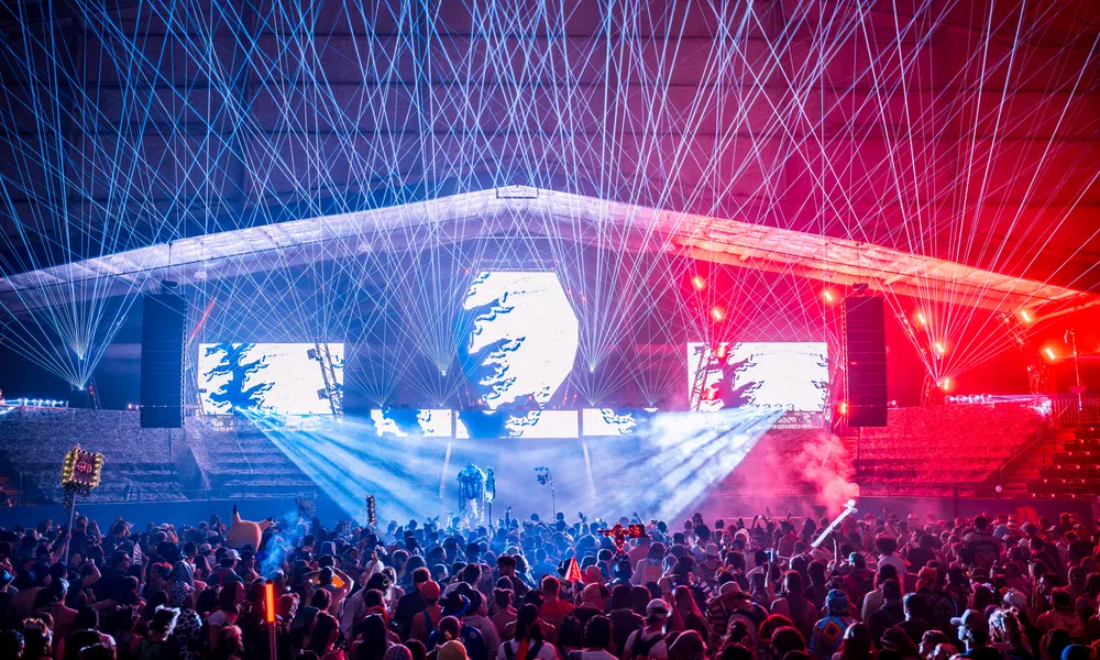 Crowd enjoying a nighttime concert with vibrant laser lights and colorful stage effects under a large canopy.