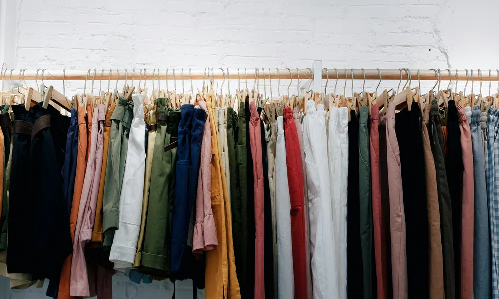Variety of colorful pants and trousers hanging on a wooden rack against a white brick wall.