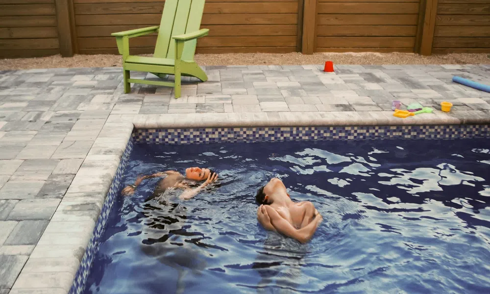 Two boys swimming and playing in a backyard pool next to a green Adirondack chair on a stone patio.