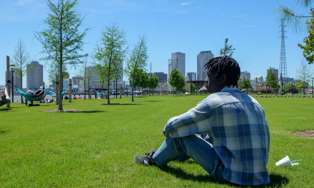 Young man in plaid shirt sitting on green grass with city skyline and trees in background on clear day
