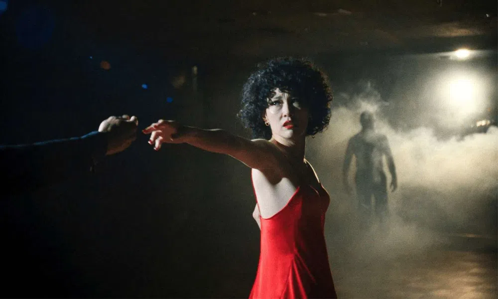 Woman in a red dress reaching out hand in a dark room with mist and a shadowy figure in the background