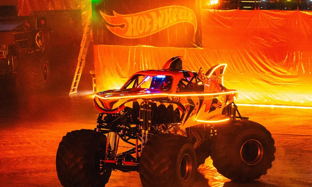 Monster truck with tiger stripes and glowing accents performs indoors in front of Hot Wheels banner at night.