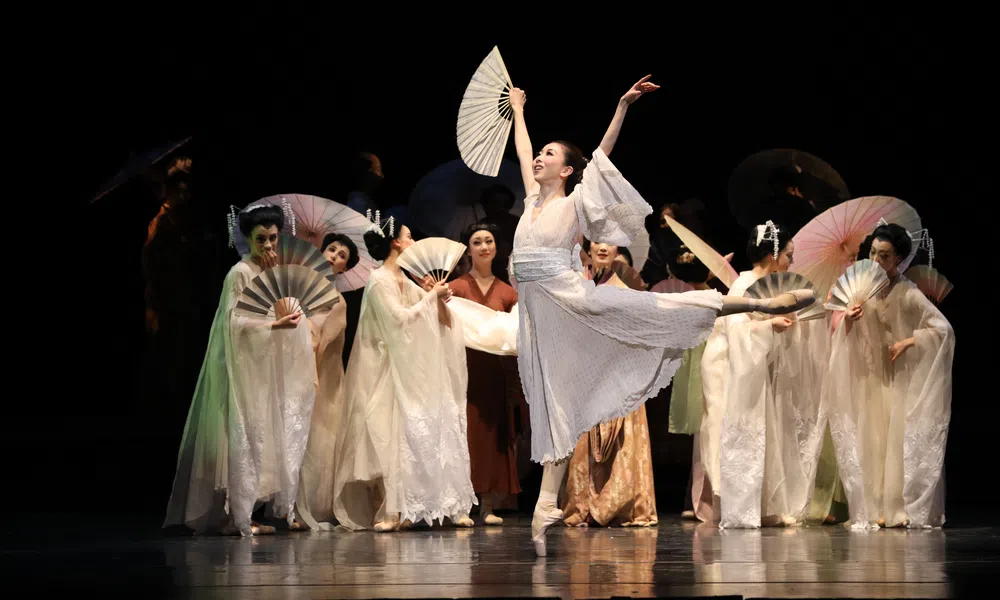 Ballet dancer in white costume performs with fans on stage with other dancers in traditional attire.