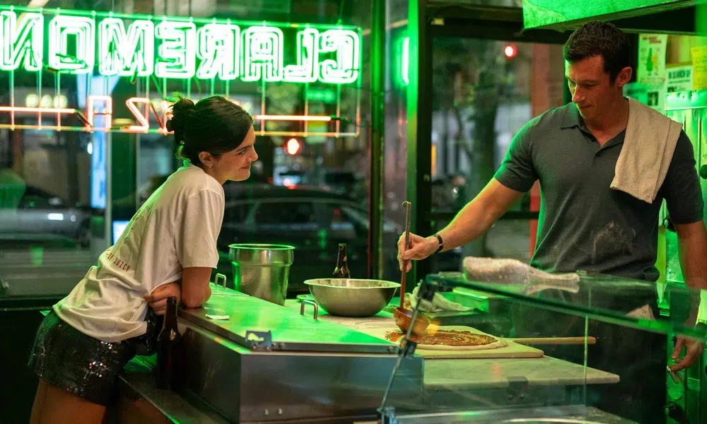 Man preparing pizza behind counter while woman watches inside a neon-lit pizzeria at night.