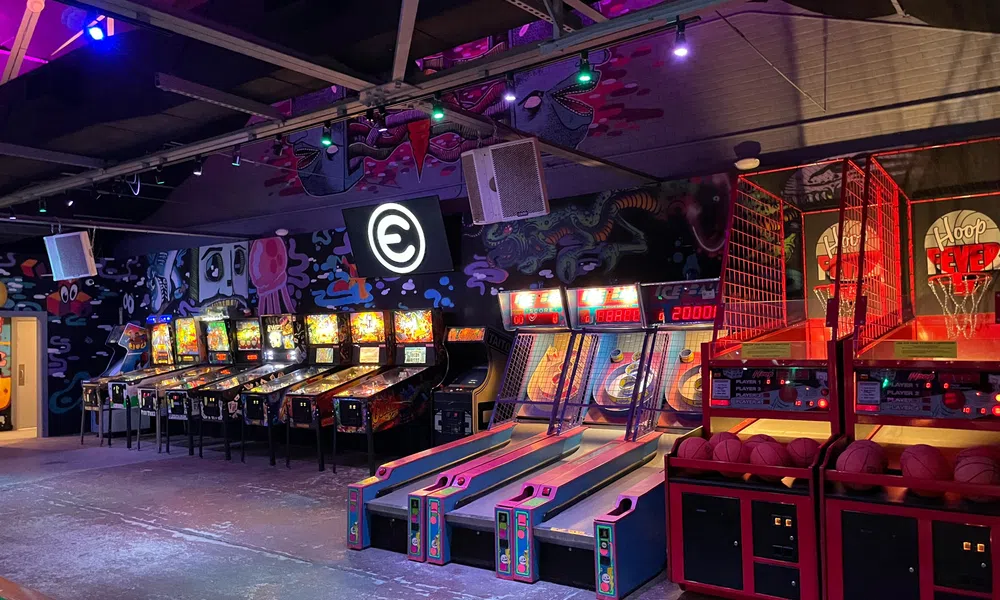 Indoor arcade with pinball machines, skee ball lanes, and basketball hoops under neon lighting.