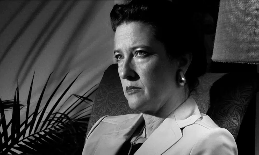 Black and white portrait of a serious woman sitting in a chair with shadows and a lamp in the background