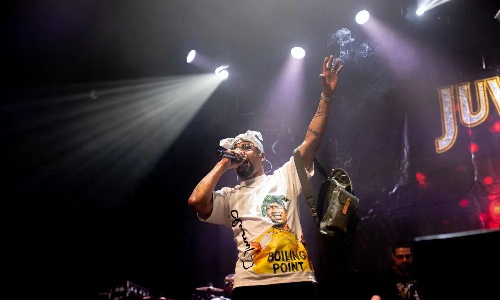 Hip hop artist performing on stage under bright spotlights with microphone and raised arm during concert.