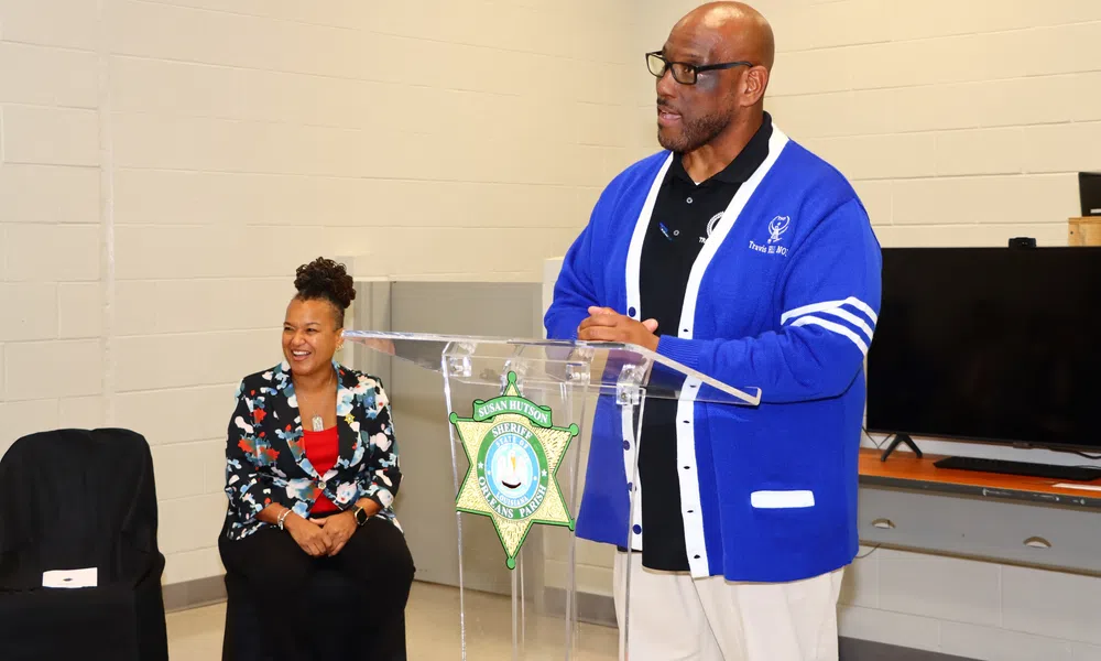 Man in blue cardigan speaking at podium with sheriff badge, woman in floral blazer seated nearby smiling