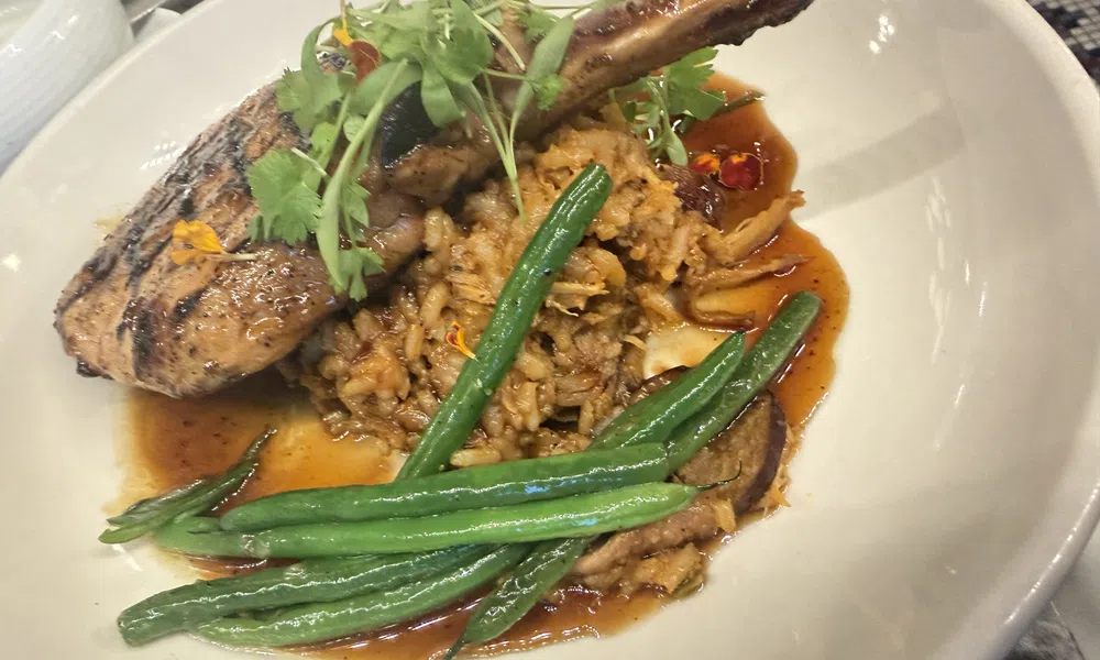 Grilled pork chop with green beans, rice, and sauce garnished with fresh herbs in a white bowl.
