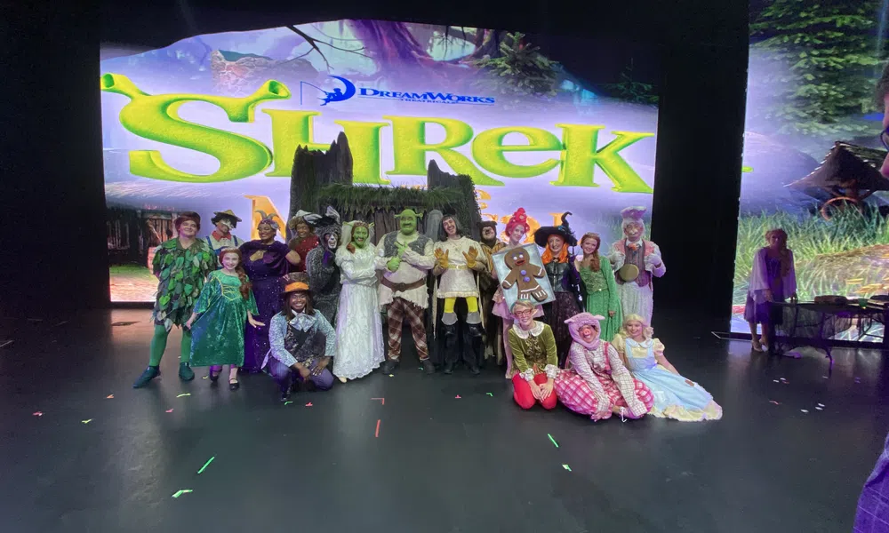 Cast of Shrek musical in full costumes posing on stage with Shrek show title backdrop