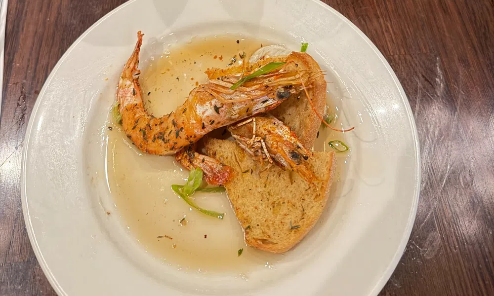 Two grilled shrimp served on toasted bread with broth and garnished with herbs on a white plate.