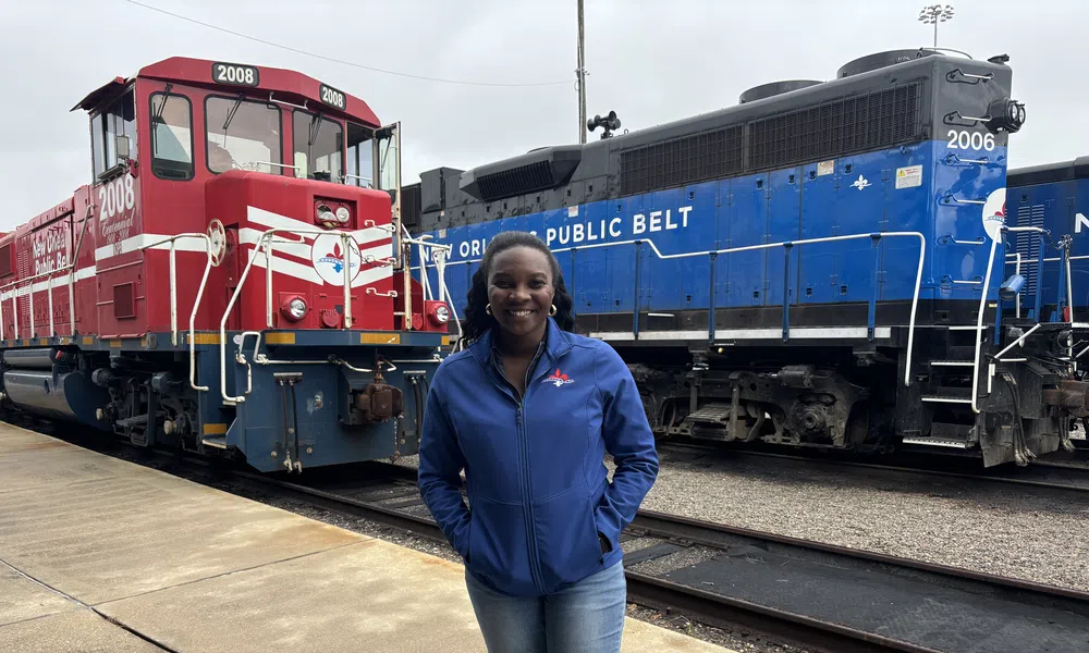 Smiling woman in blue jacket standing on platform between red and blue trains at a railway yard.