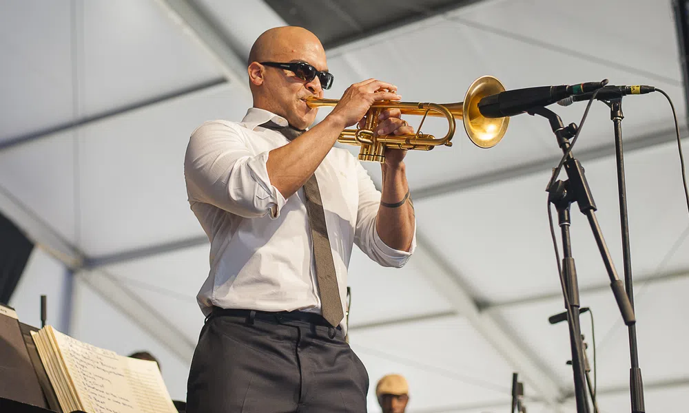 Bald man wearing sunglasses playing trumpet on stage under white tent with microphone nearby