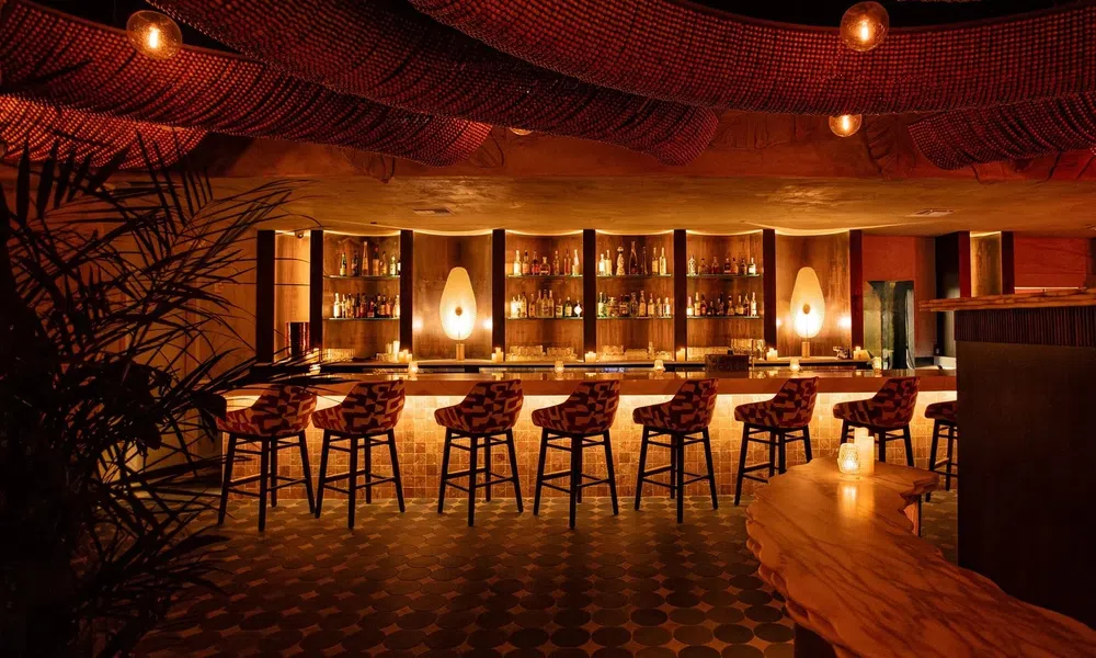 Cozy dimly lit bar with wooden stools, patterned ceiling, and warm ambient lighting over the liquor shelves.