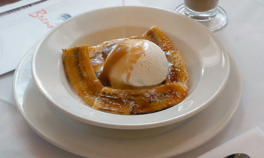 Grilled plantains topped with vanilla ice cream and caramel sauce in a white bowl on a white table.