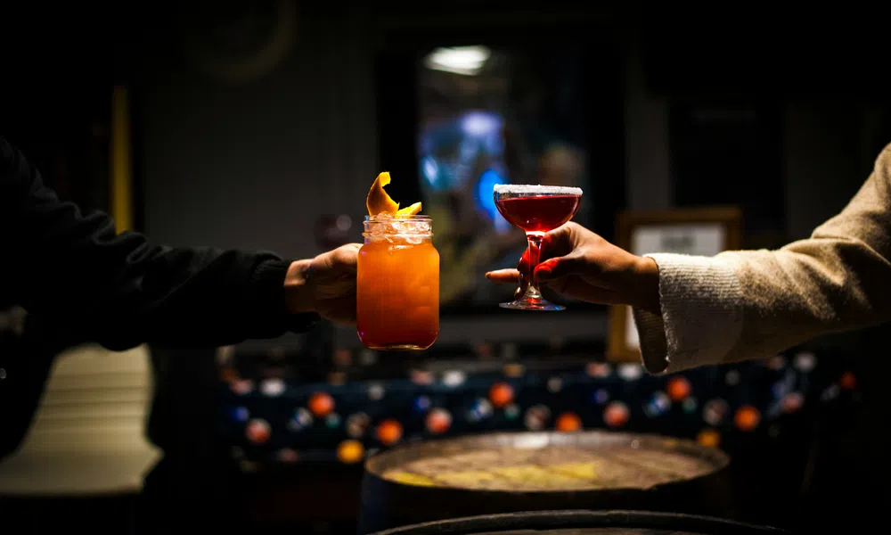 Two hands holding up colorful cocktails in dimly lit bar with wooden barrels in the foreground.