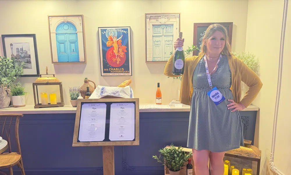 Woman holding wine bottle inside cozy wine bar with framed art, menu stand, plants, and candles in background.