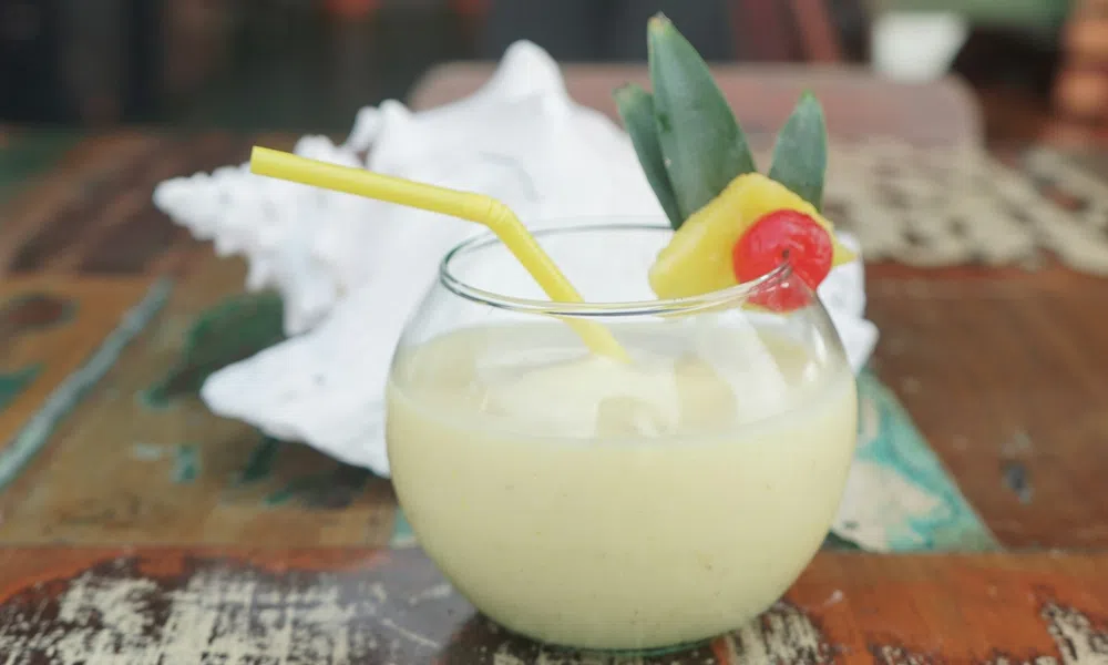 Pineapple smoothie in a round glass with a yellow straw, garnished with pineapple slice and cherry on a rustic wooden table.