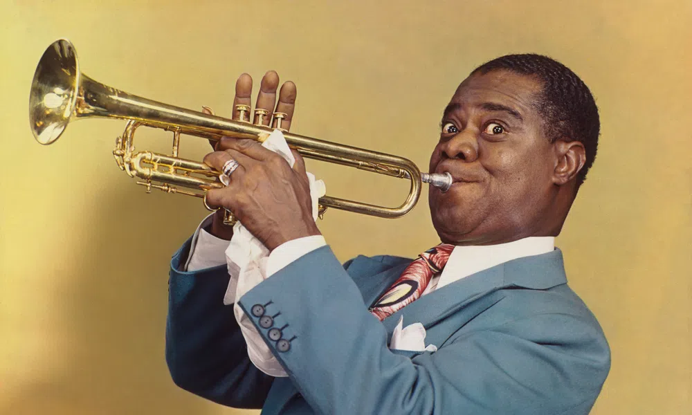 Man in blue suit playing trumpet with focused expression on yellow background.