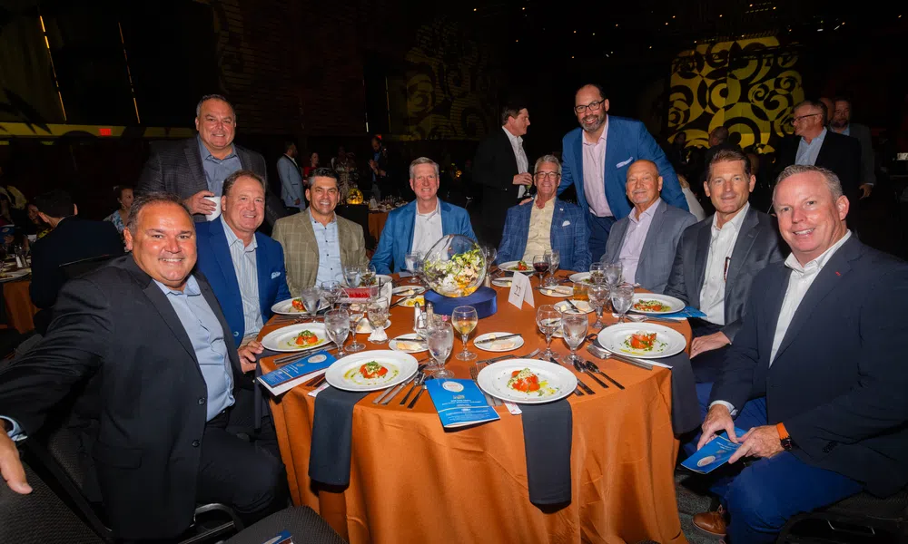 Group of men in suits gathered around a round table at a formal dinner event with plated food and drinks.
