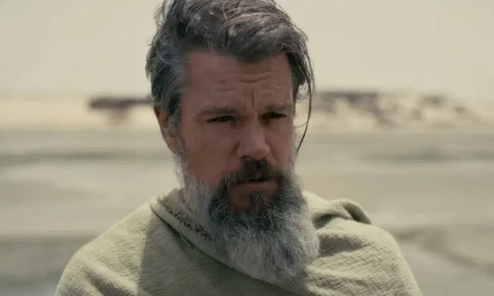Man with gray beard and hair wearing a green cloak in a desert landscape with blurred sandy background.
