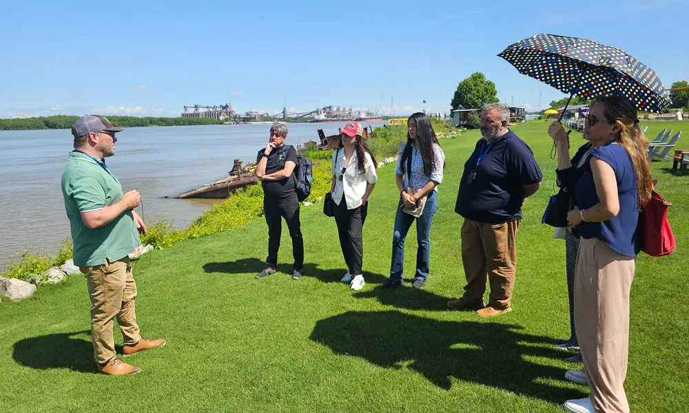 Group of six adults listening to a man speaking by a riverside on a sunny day with clear blue sky.