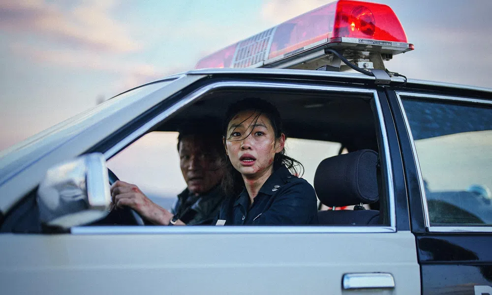Female police officer and male colleague in a police car with red siren light, looking concerned through open window.