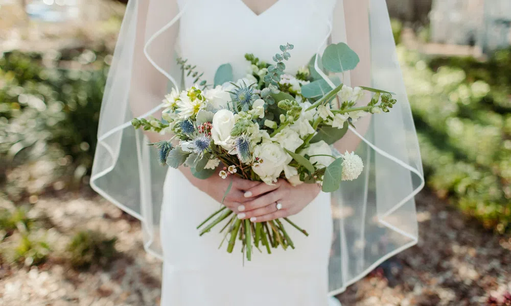 Bride in white dress holding bouquet of white and green flowers with veil outdoors on sunny day