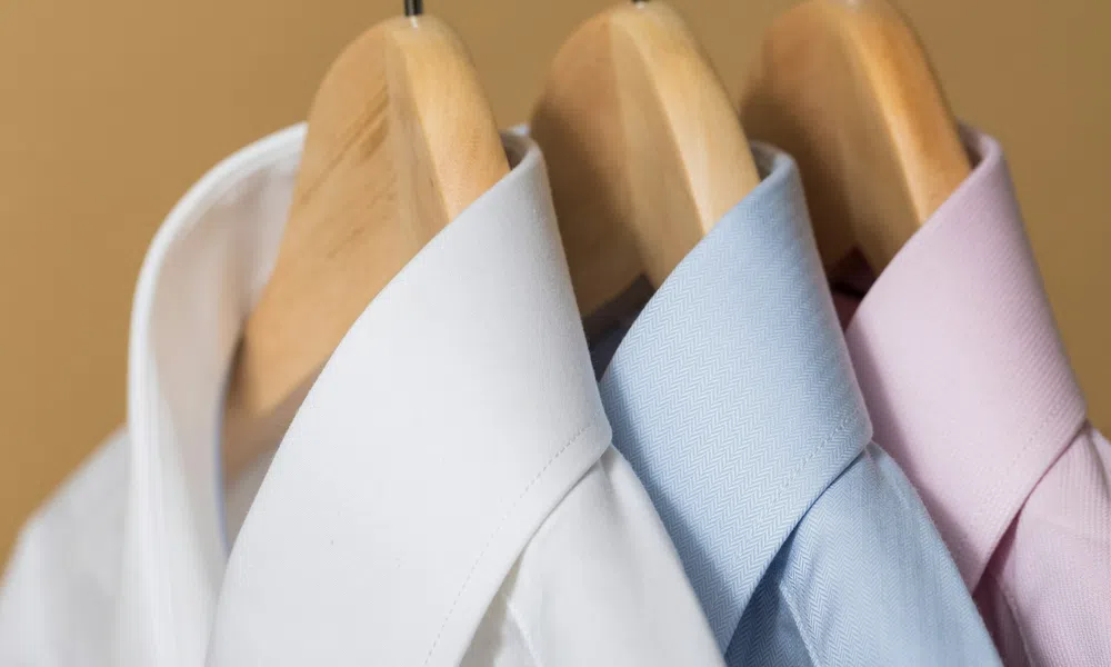 White, blue, and pink dress shirts neatly hanging on wooden hangers against a beige background