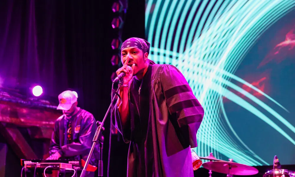 Hip-hop artist performing on stage with microphone and DJ in the background under vibrant purple and blue lights.