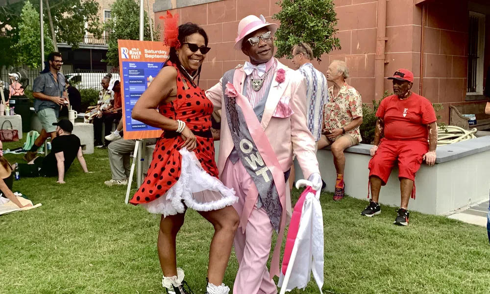 Couple dressed in vibrant retro outfits enjoying an outdoor festival on a sunny day with people relaxing nearby
