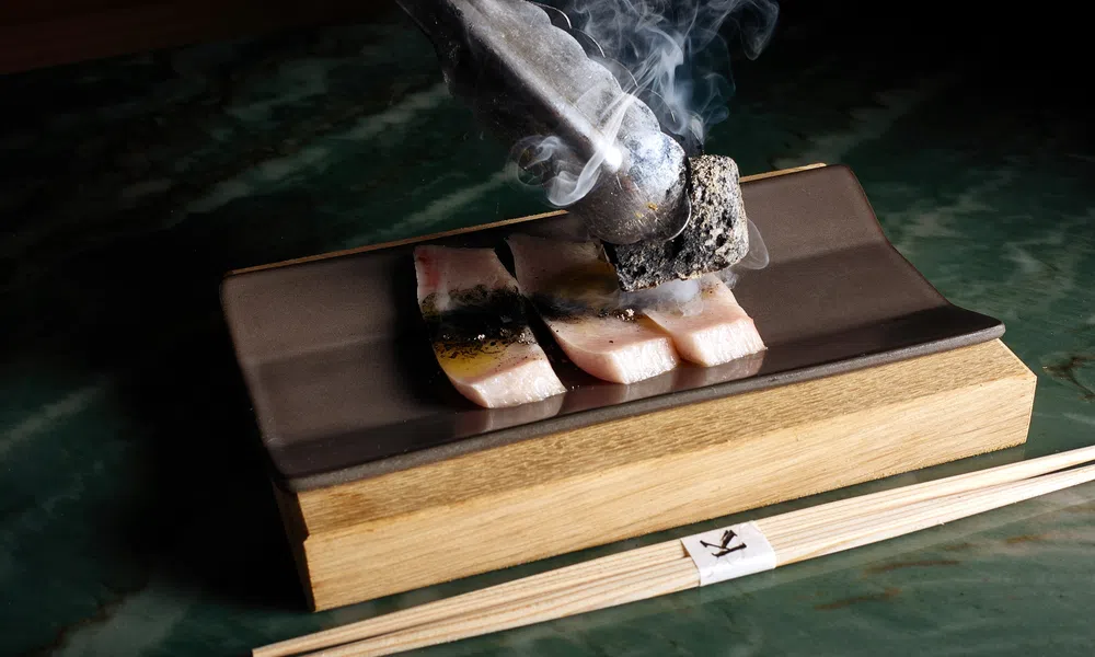 Close-up of sizzling fish slices being grilled with smoking charcoal on a rectangular plate with chopsticks.