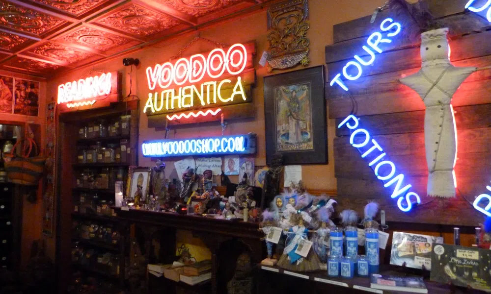 Interior of a voodoo shop with neon signs, shelves of jars, potions, dolls, and mystical items under ornate ceiling tiles.