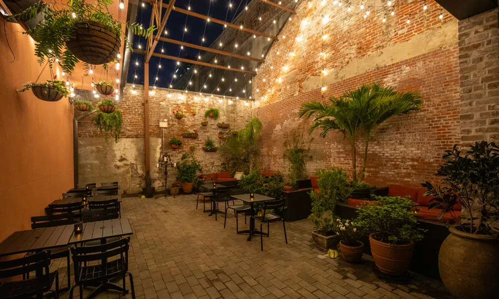 Cozy outdoor dining area with string lights, brick walls, potted plants, and black tables and chairs at night.