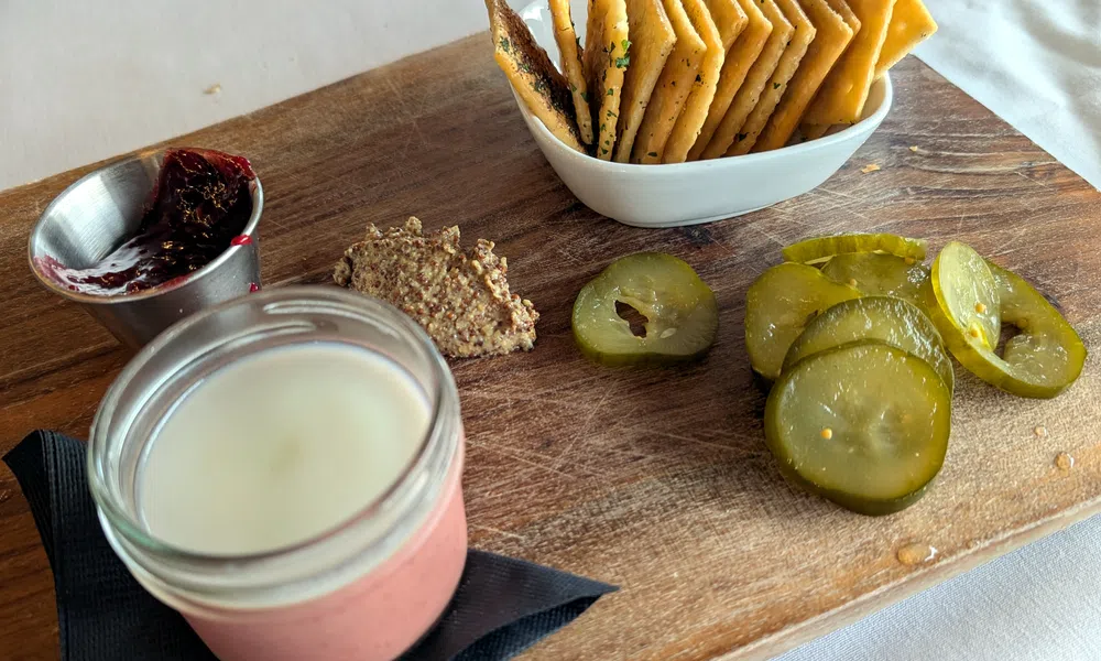 Charcuterie board with crackers, sliced pickles, mustard, berry jam, and a jar of pâté with creamy topping.
