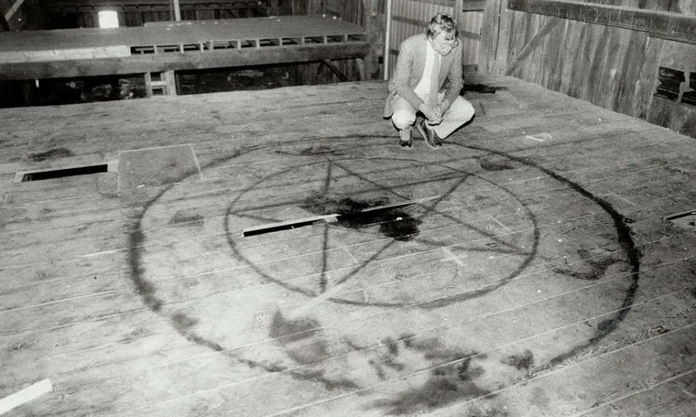 Black and white image of a man inspecting a large pentagram symbol drawn on a wooden floor inside a barn.