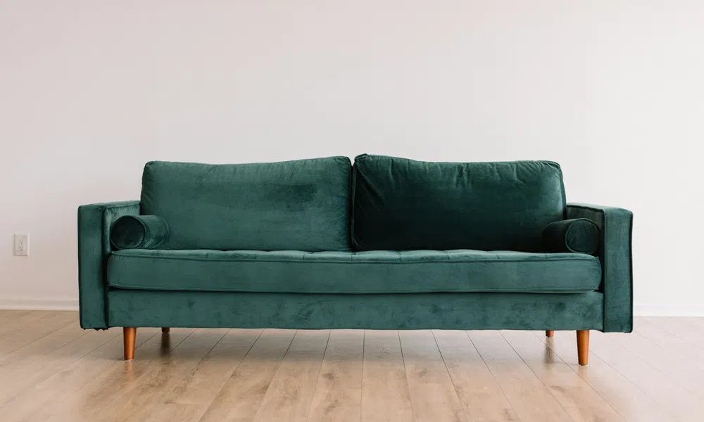 Modern emerald green velvet sofa with wooden legs on light wooden floor against plain white wall