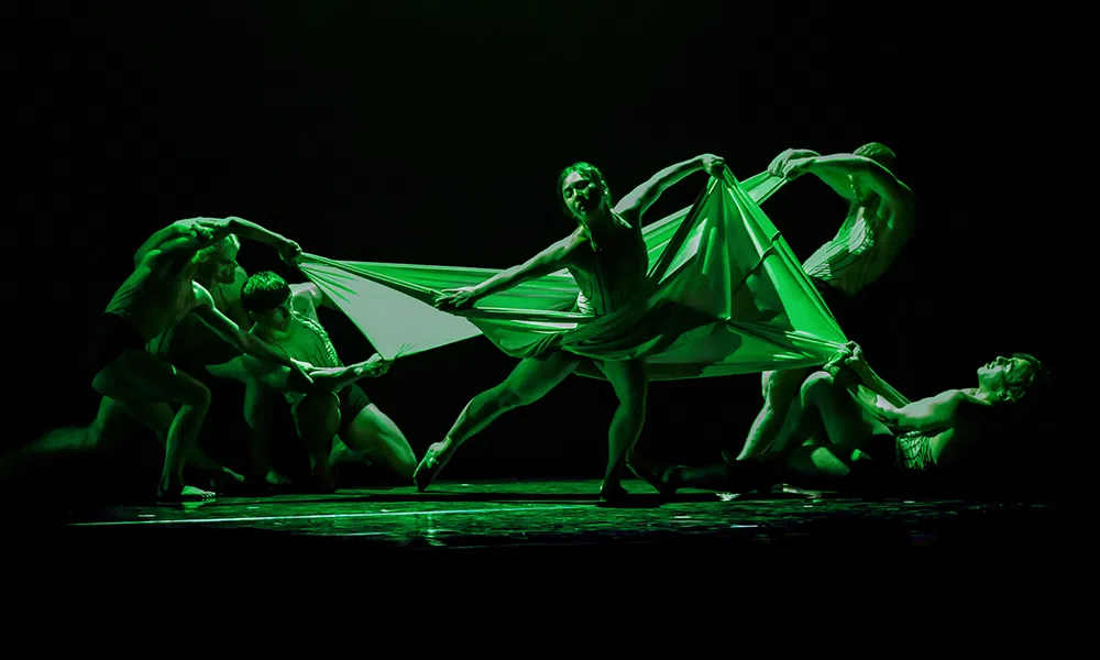 Performers in a dramatic green-lit dance holding and stretching a large fabric on stage in dynamic poses.