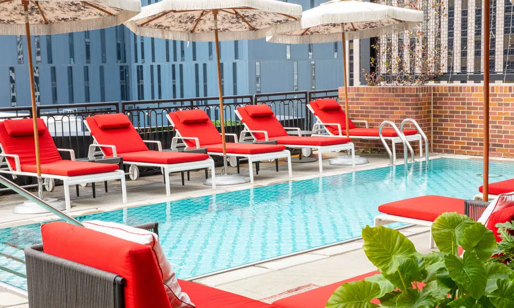 Outdoor pool area with red lounge chairs and umbrellas on a sunny day, surrounded by urban buildings.