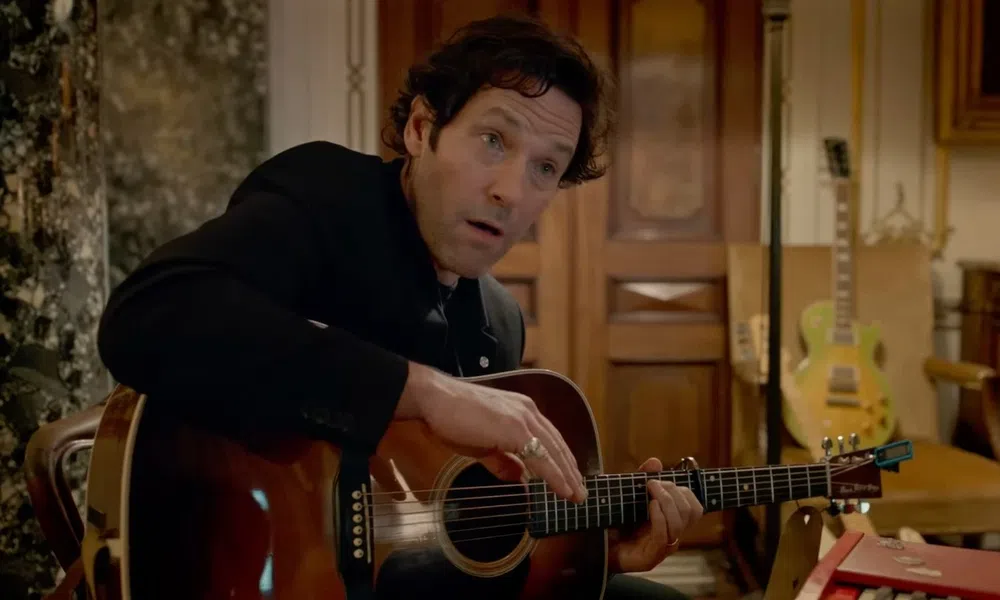 Man in black jacket playing acoustic guitar indoors with wooden doors and electric guitar in background