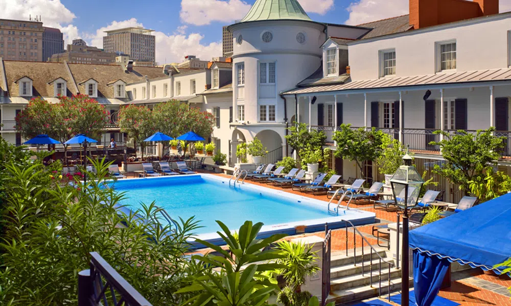 Outdoor pool area surrounded by lounge chairs, greenery, and historic white buildings under a blue sky.