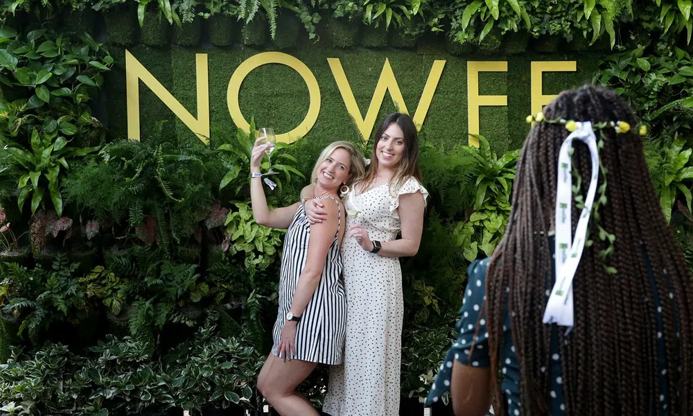 Two women pose and smile in front of a green plant wall with large yellow NOWEF letters while another person photographs them.