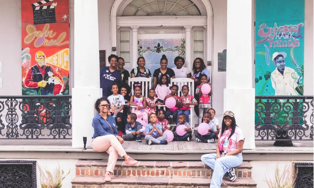 Group of adults and children posing with pink balloons on porch with colorful artwork on walls