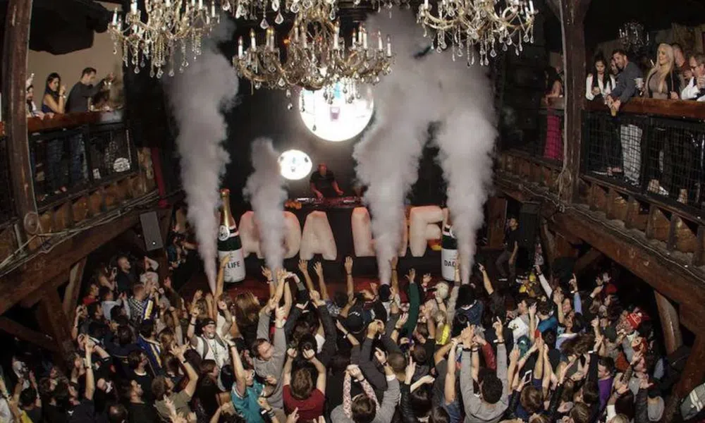 Crowded nightclub with people celebrating under chandeliers and large smoke effects from bottles on the dance floor.
