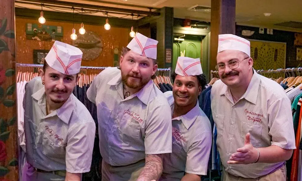 Four men wearing retro diner uniforms and hats posing with outstretched hands in a cozy restaurant setting.