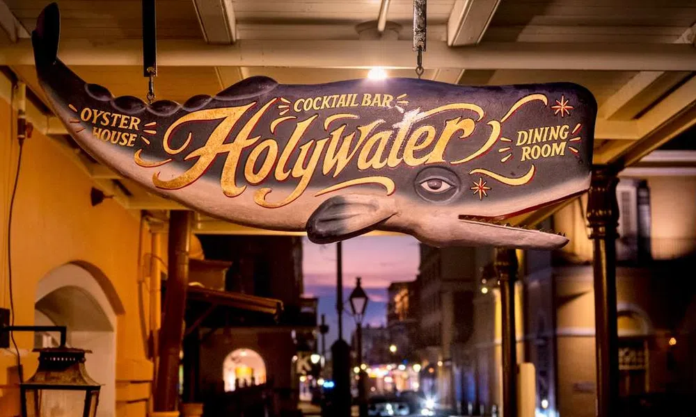 Hanging whale-shaped wooden sign for Holy Water oyster house, cocktail bar, and dining room at dusk.