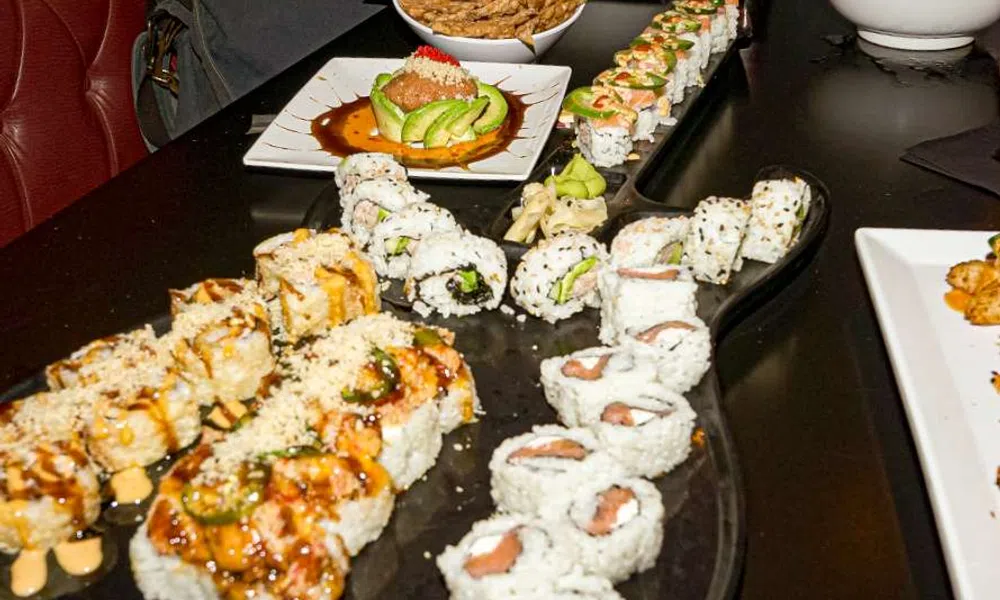 Variety of sushi rolls arranged on a black table with sauces, avocado, and garnishes in a restaurant setting.